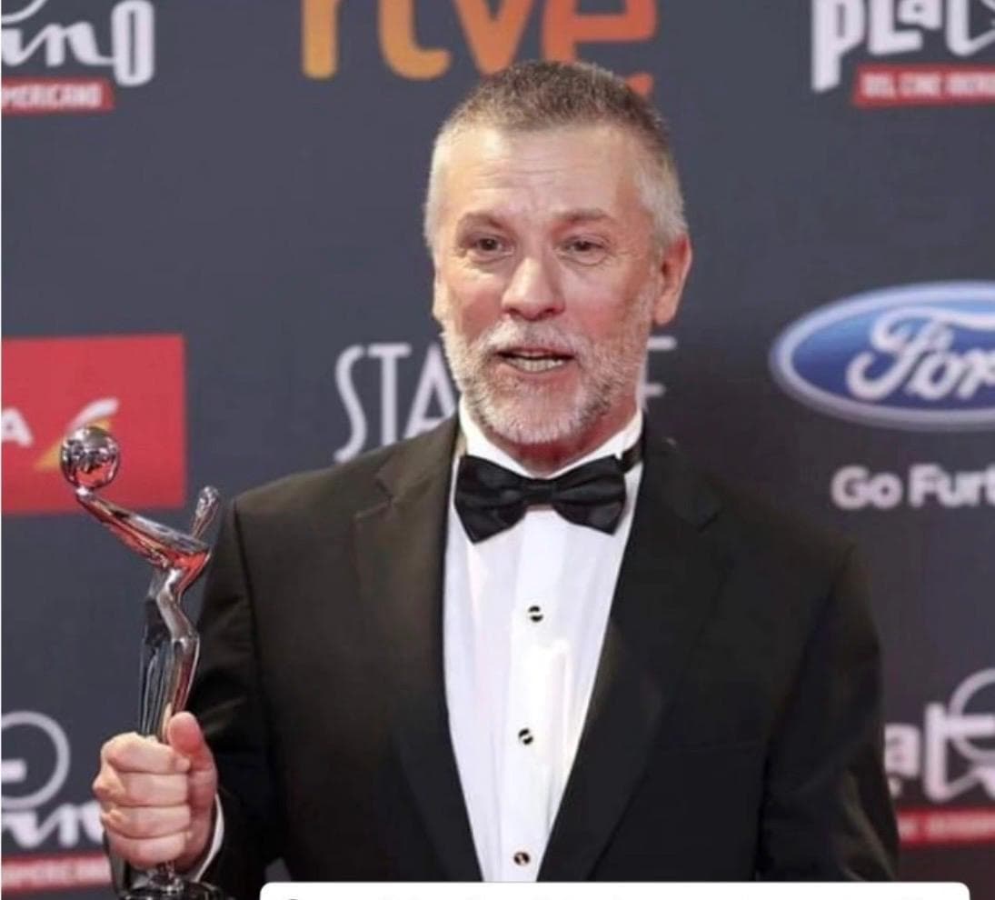 José Luis Díaz con esmoquin y moño, sosteniendo un trofeo de los Premios Platino del Cine Iberoamericano, ante un fondo del photocall del evento.